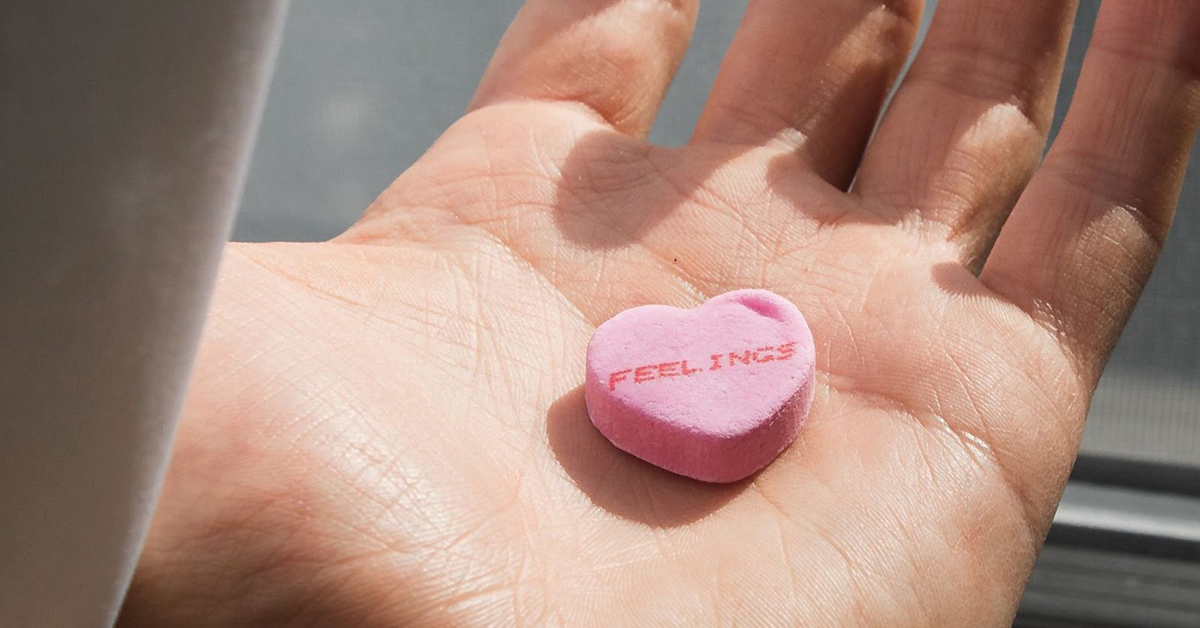 Main qui tient un bonbon rose en forme de cœur où il est inscrit "feeling"
